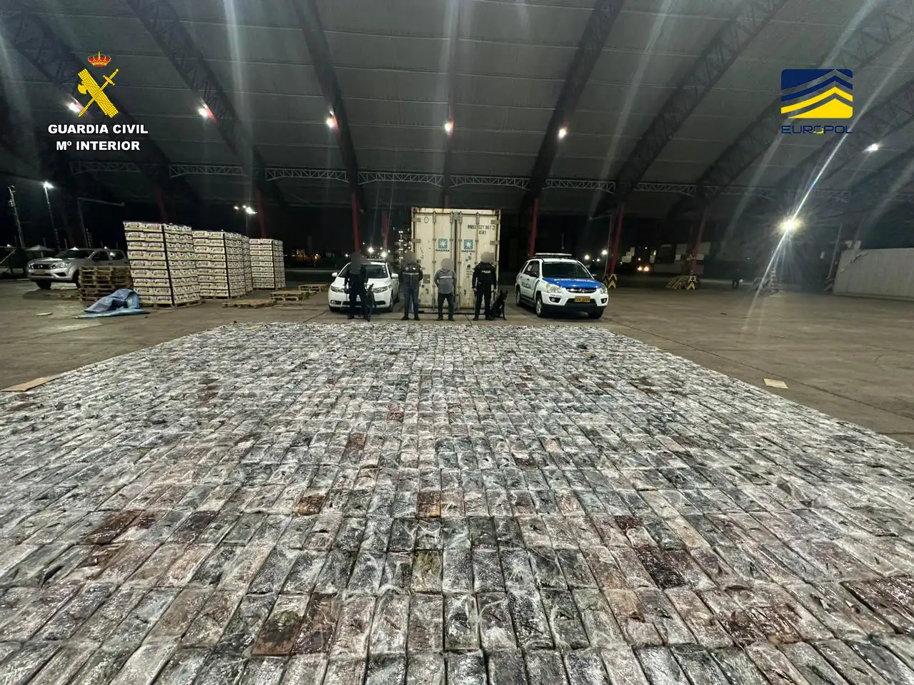 Cientos de paquetes de droga plastificados extendidos en el suelo de una nave portuaria de noche; al fondo, un contenedor frigorífico, dos coches policiales y varios agentes con el rostro pixelado junto a un perro detector.