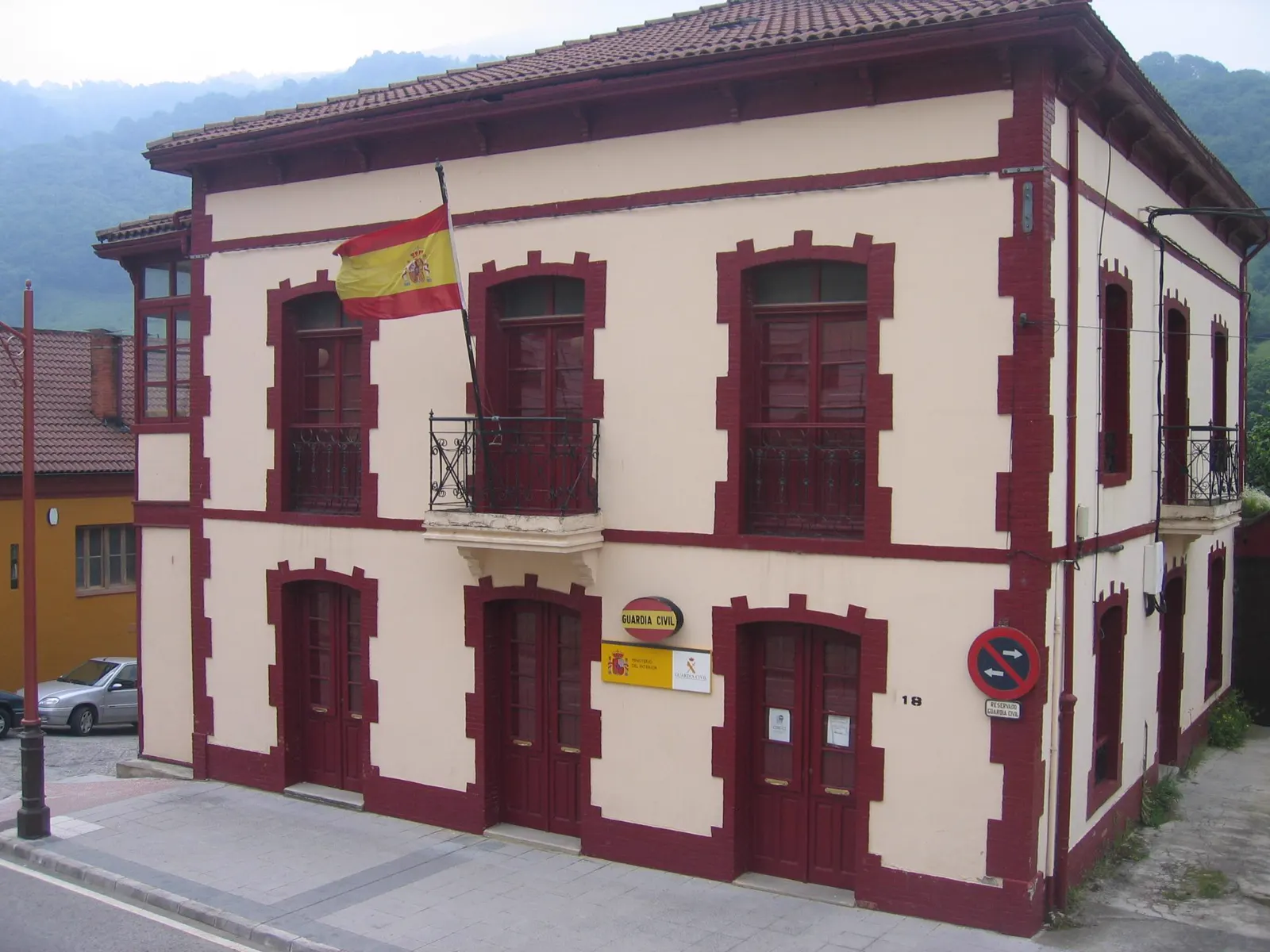 Fachada de un edificio de dos plantas encalado, con recercados de ladrillo rojo, bandera de España en el balcón y un cartel con el emblema de la Guardia Civil junto a la puerta