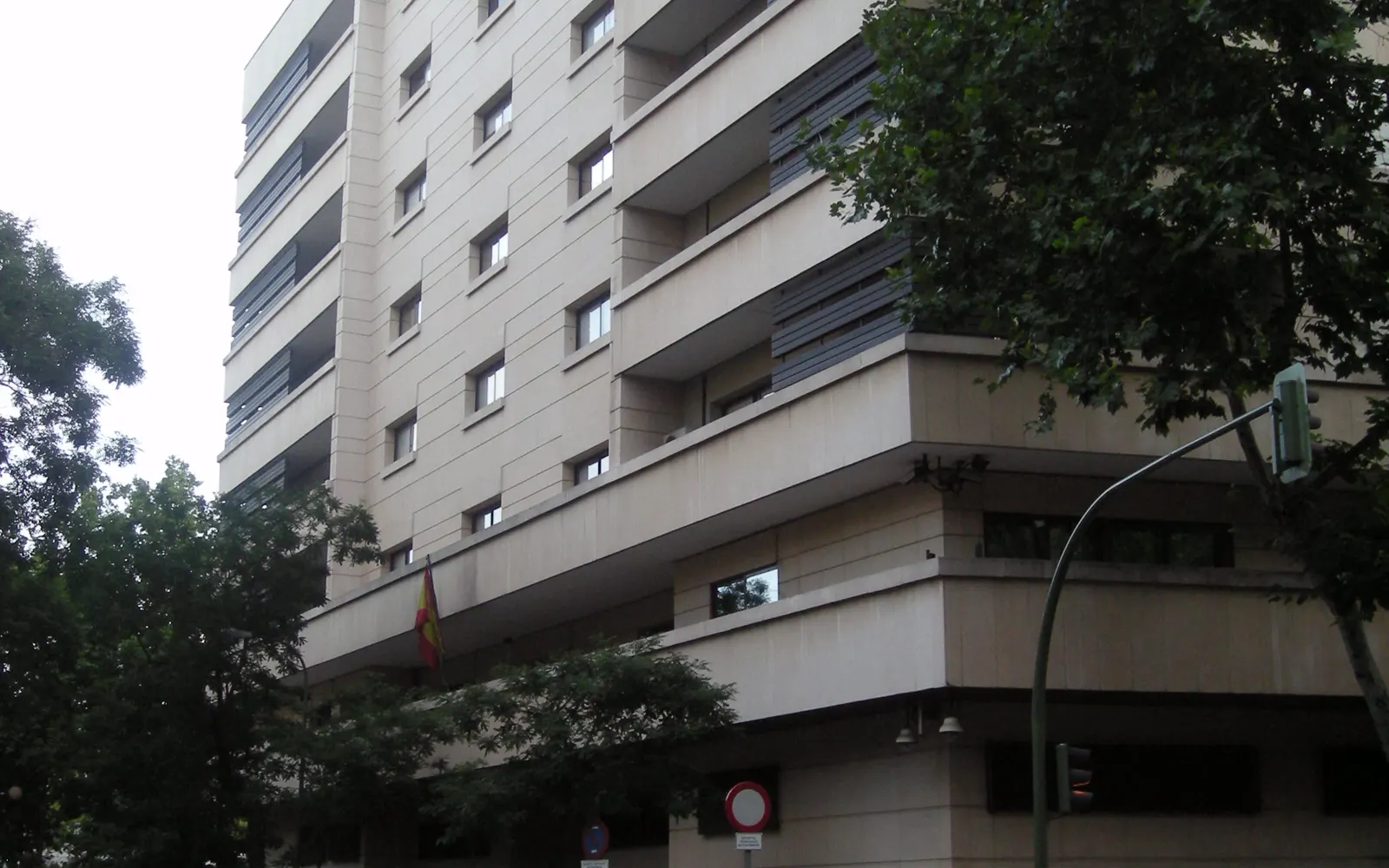 Fachada de una sede de la Audiencia Nacional en Madrid: edificio de piedra clara con voladizos horizontales y ventanas estrechas, una bandera de España colgada sobre la entrada y árboles delante.