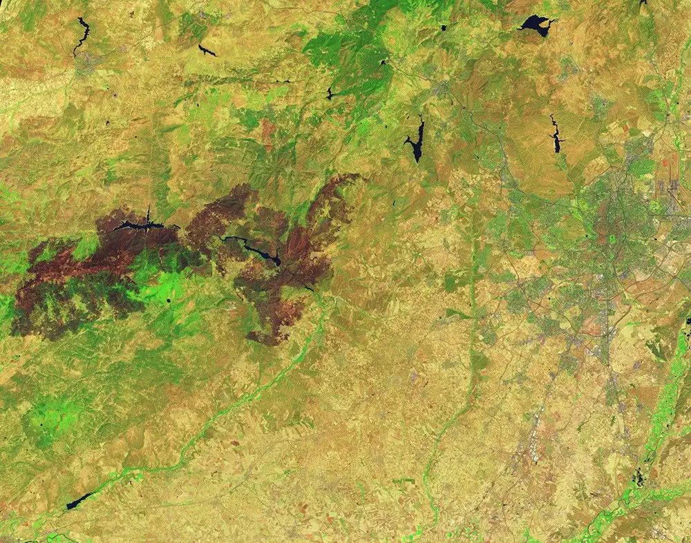 Imagen de satélite en falso color del centro peninsular: sobre un mosaico de tonos ocres y verdes se extiende de este a oeste una gran mancha marrón oscura de vegetación quemada, atravesada por dos embalses alargados de agua negra; a la derecha se distingue la mancha gris de la aglomeración de Madrid con sus carreteras radiales.