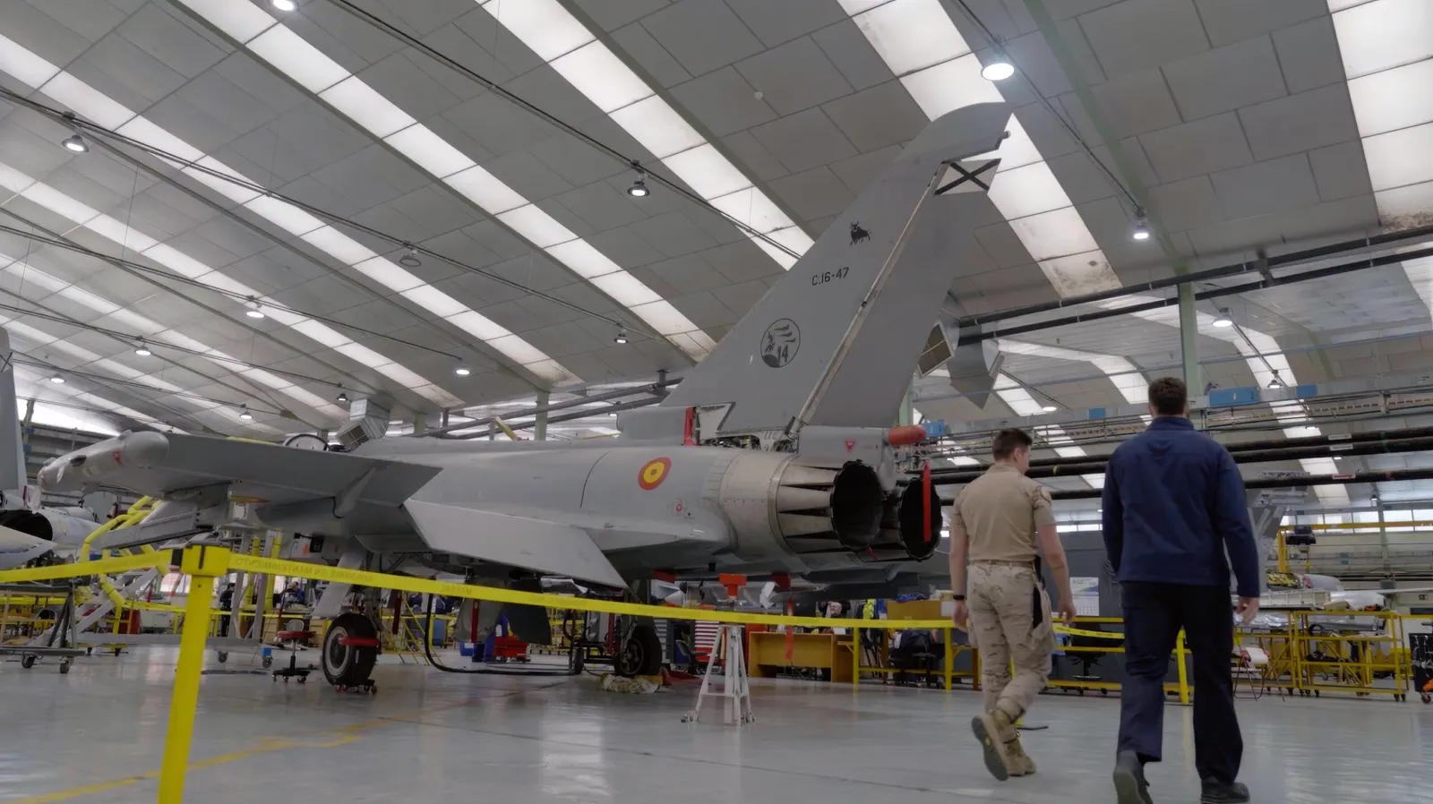 Un militar con uniforme de campaña y un técnico con mono azul caminan hacia un caza aparcado en un hangar de mantenimiento, con otras aeronaves desmontadas al fondo.