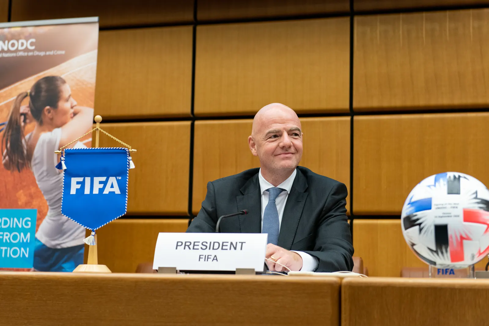 Gianni Infantino, con traje oscuro y corbata azul, sentado y sonriente tras una mesa con un cartel que dice PRESIDENT FIFA, un banderín de la FIFA a su izquierda y un balón sobre un pedestal a su derecha