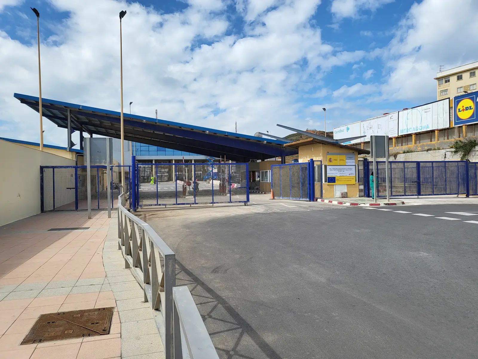 Puesto fronterizo de El Tarajal, en Ceuta: una marquesina metálica azul sobre los controles, vallas azules cerradas, la calzada vacía y, al fondo, una ladera con edificios y vallas publicitarias bajo un cielo con nubes.