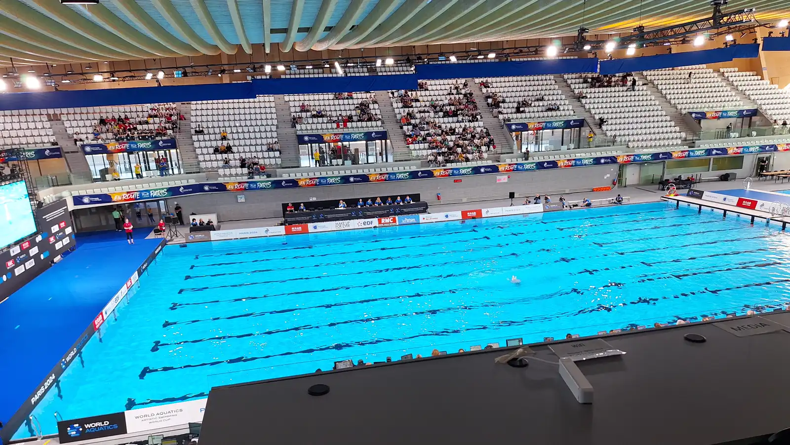 Vista general de la piscina de competición del Centro Acuático Olímpico de Saint-Denis, con el techo de madera curvado, el graderío blanco a medio llenar, la mesa de jueces junto al vaso y un cartel de la Copa del Mundo de natación artística de World Aquatics al fondo.