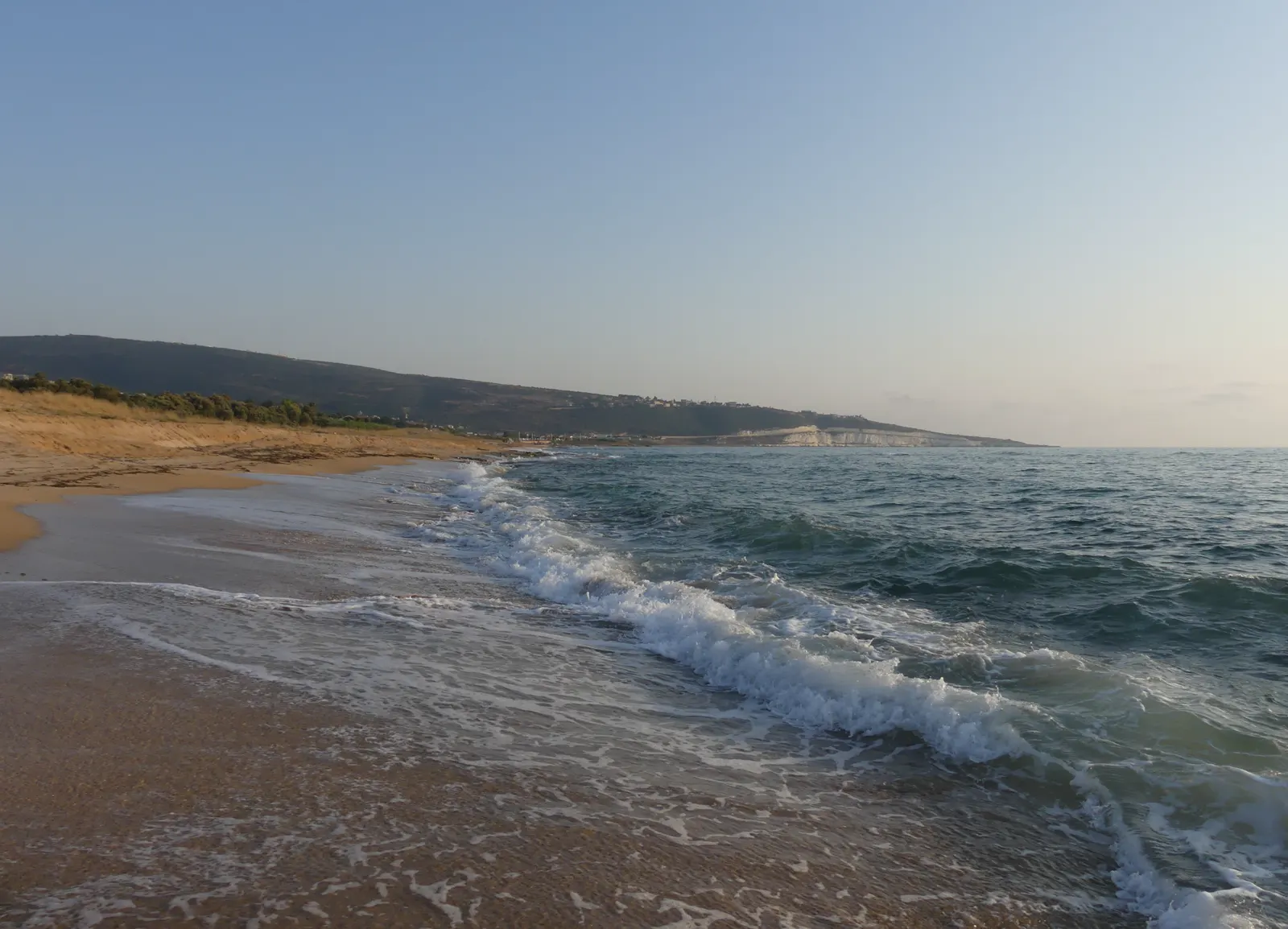 Playa de arena y rompiente del mar en Mansuri, con una loma verde y casas dispersas al fondo bajo un cielo despejado