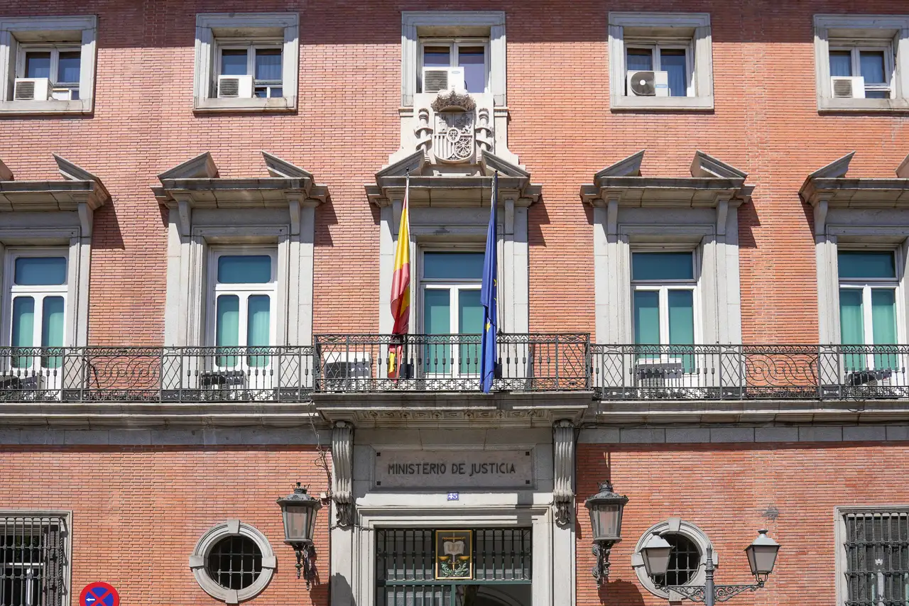 Fachada de ladrillo rojo del Ministerio de Justicia en Madrid, con balcones de forja, el escudo real tallado en piedra sobre el balcón central, las banderas de España y de la Unión Europea y el rótulo «Ministerio de Justicia» grabado sobre la puerta principal.