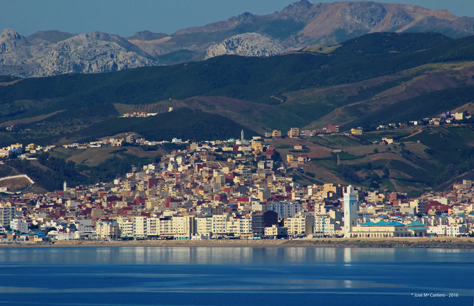 Vista de la ciudad marroquí de Fnideq (Castillejos) desde Ceuta, al otro lado del agua: una franja de edificios blancos junto a la playa, una mezquita con minarete azul y blanco a la derecha, casas de colores trepando por la ladera y las montañas del Rif al fondo.