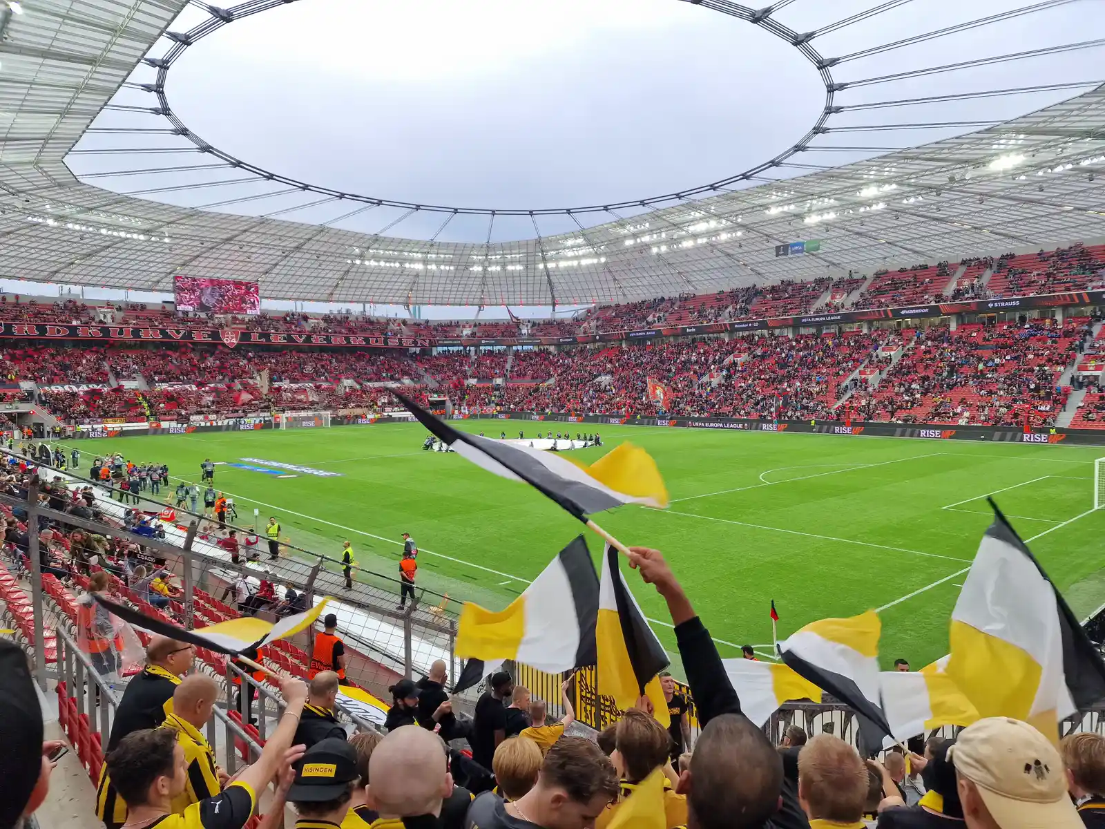 Interior del estadio BayArena de Leverkusen lleno de público durante un partido nocturno, con aficionados ondeando banderas amarillas y negras en primer plano desde la grada.