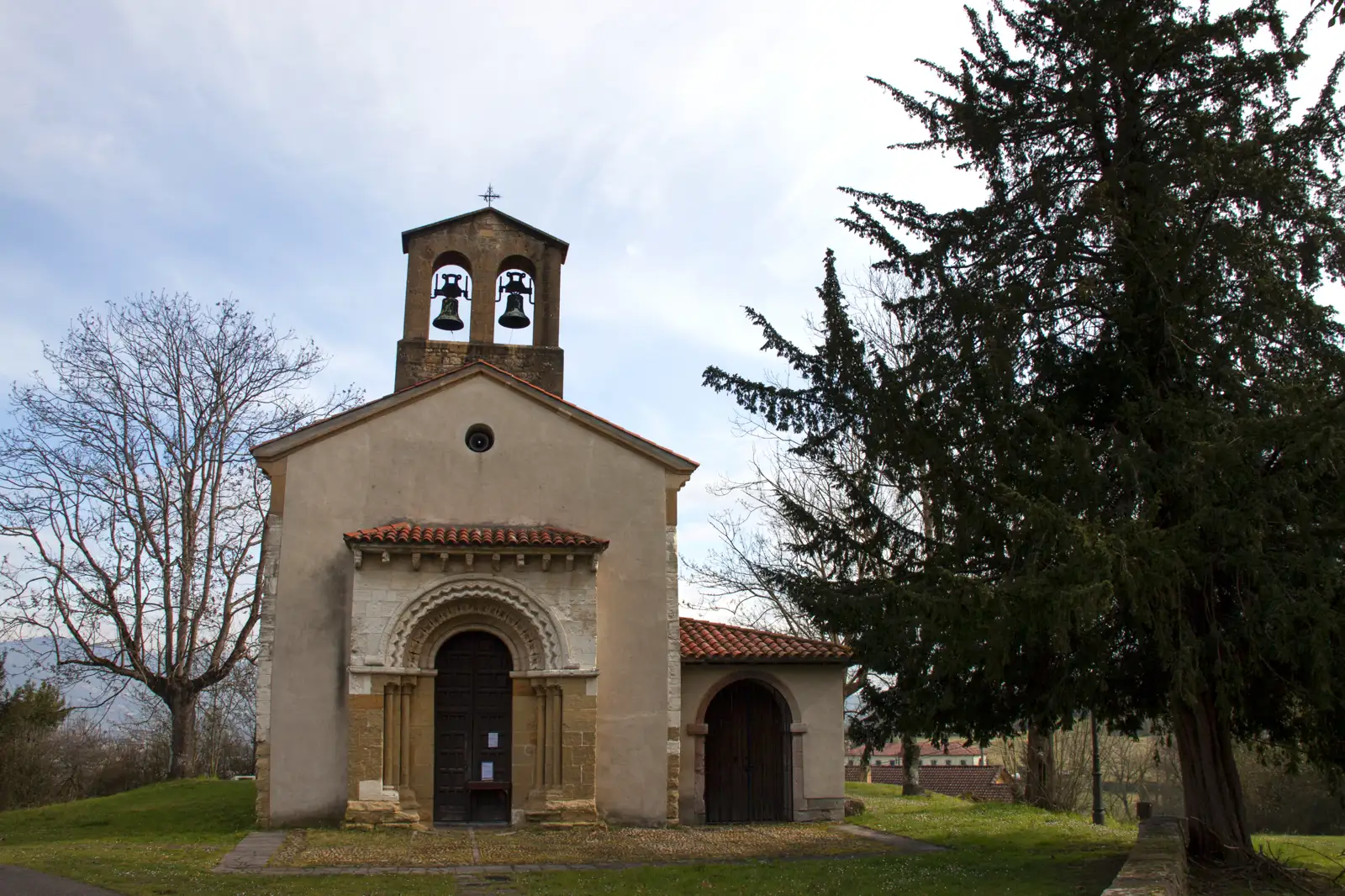 Iglesia rural de muros encalados y sillería de piedra, con espadaña de dos campanas y un pórtico de arcos, rodeada de prado, un tejo y árboles sin hoja.