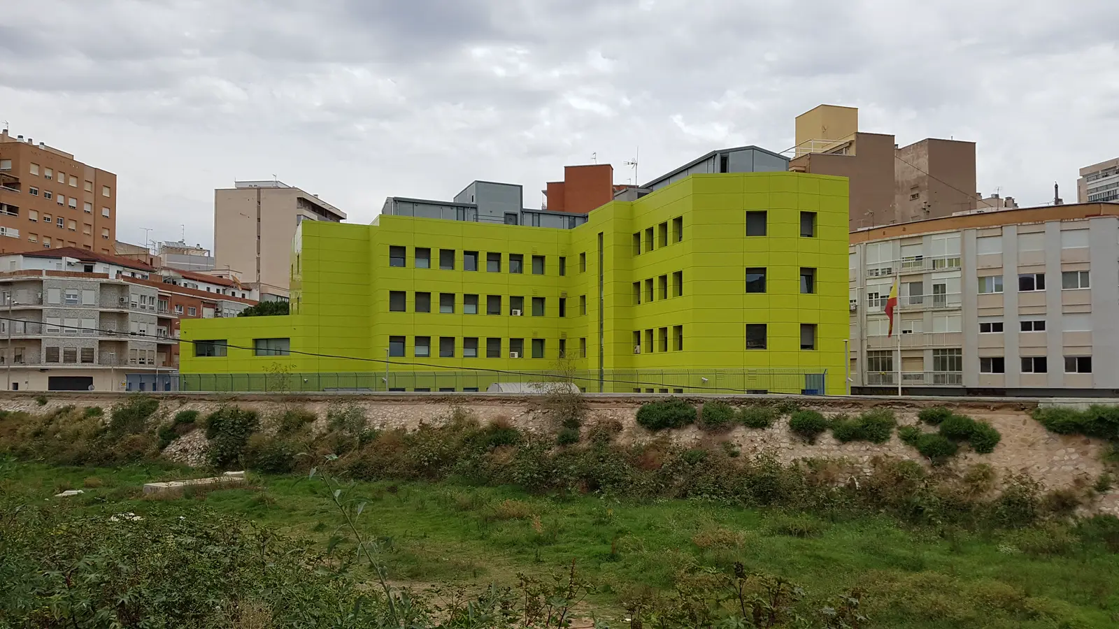 Edificio institucional de cuatro plantas con la fachada revestida en verde lima, encajado entre bloques de viviendas de una ciudad, visto desde un solar con vegetación
