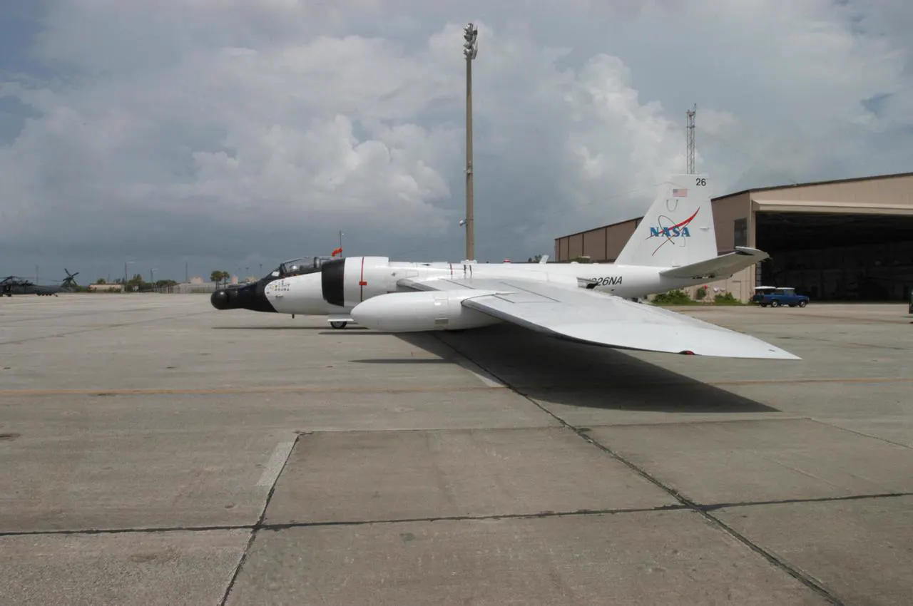 Avión de investigación WB-57 de la NASA, blanco, con morro alargado y alas muy largas, estacionado en una pista de hormigón junto a un hangar bajo un cielo con nubes