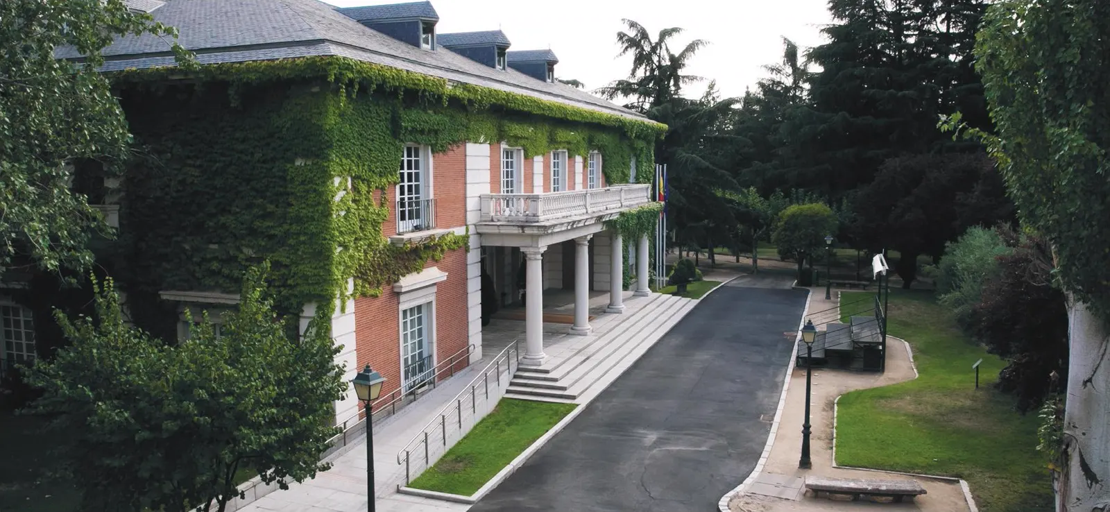 Fachada del edificio del Consejo de Ministros en el complejo de la Moncloa: construcción de ladrillo rojo cubierta de hiedra, con pórtico de columnas blancas, banderas en el balcón, y jardines y arbolado a ambos lados de la calzada de acceso.