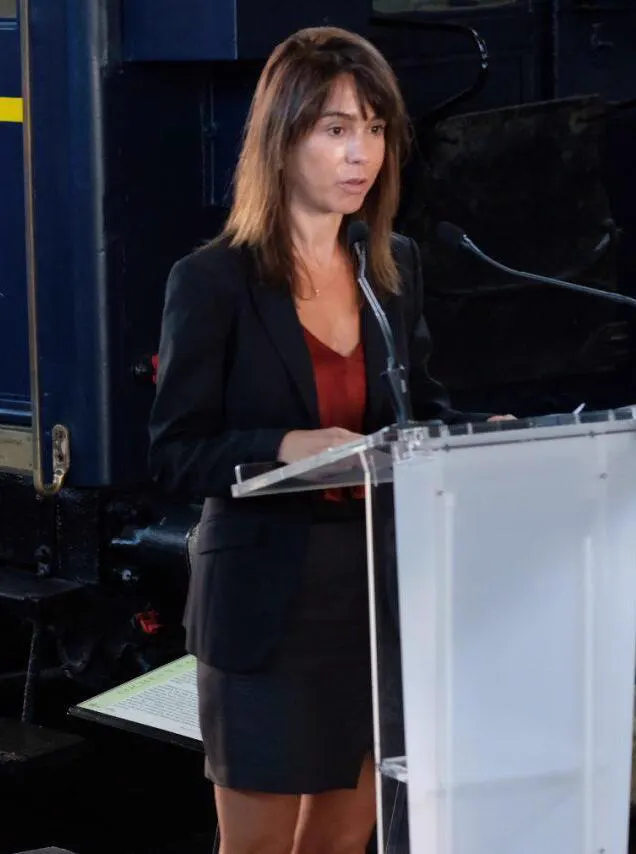 Isabel Pardo de Vera, de traje oscuro y camisa granate, habla ante un atril de metacrilato con dos micrófonos; detrás, la carrocería azul de una locomotora antigua.