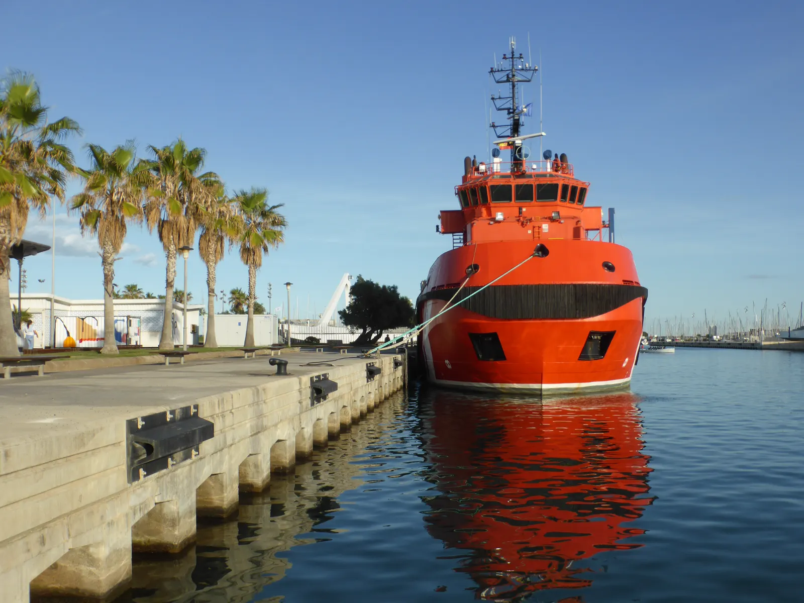 Buque de salvamento de casco naranja atracado de proa en un muelle, con palmeras a la izquierda y su reflejo en el agua
