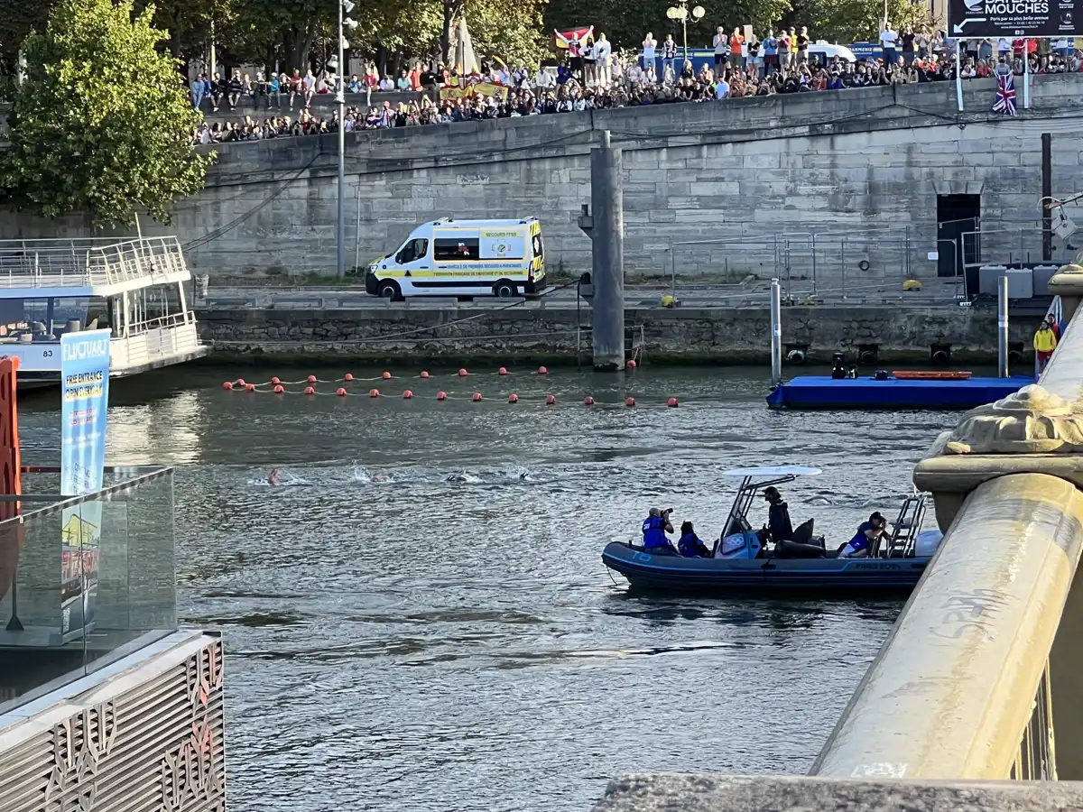 Varias nadadoras cruzan el Sena en fila junto a una línea de boyas rojas, seguidas por una lancha neumática con fotógrafos, mientras cientos de espectadores miran la carrera desde el muelle de piedra.