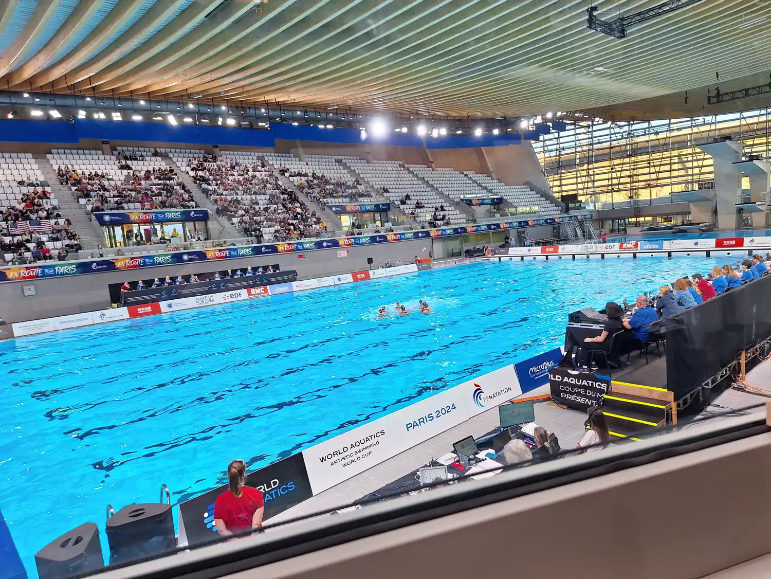 Piscina de competición del Centro Acuático Olímpico de Saint-Denis vista desde la tribuna, con nadadoras de natación artística agrupadas en el centro del vaso, el jurado sentado a la derecha del borde y un cartel de la Copa del Mundo de natación artística de World Aquatics junto al agua.