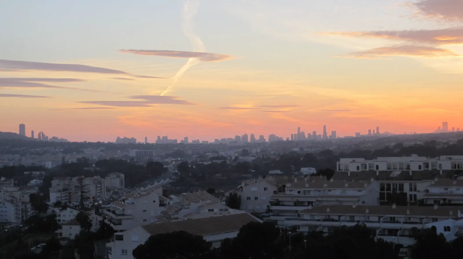 Bloques bajos de viviendas blancas con tejados de teja en primer plano y, al fondo, la silueta de las torres de apartamentos de Benidorm recortada contra un cielo anaranjado al atardecer.