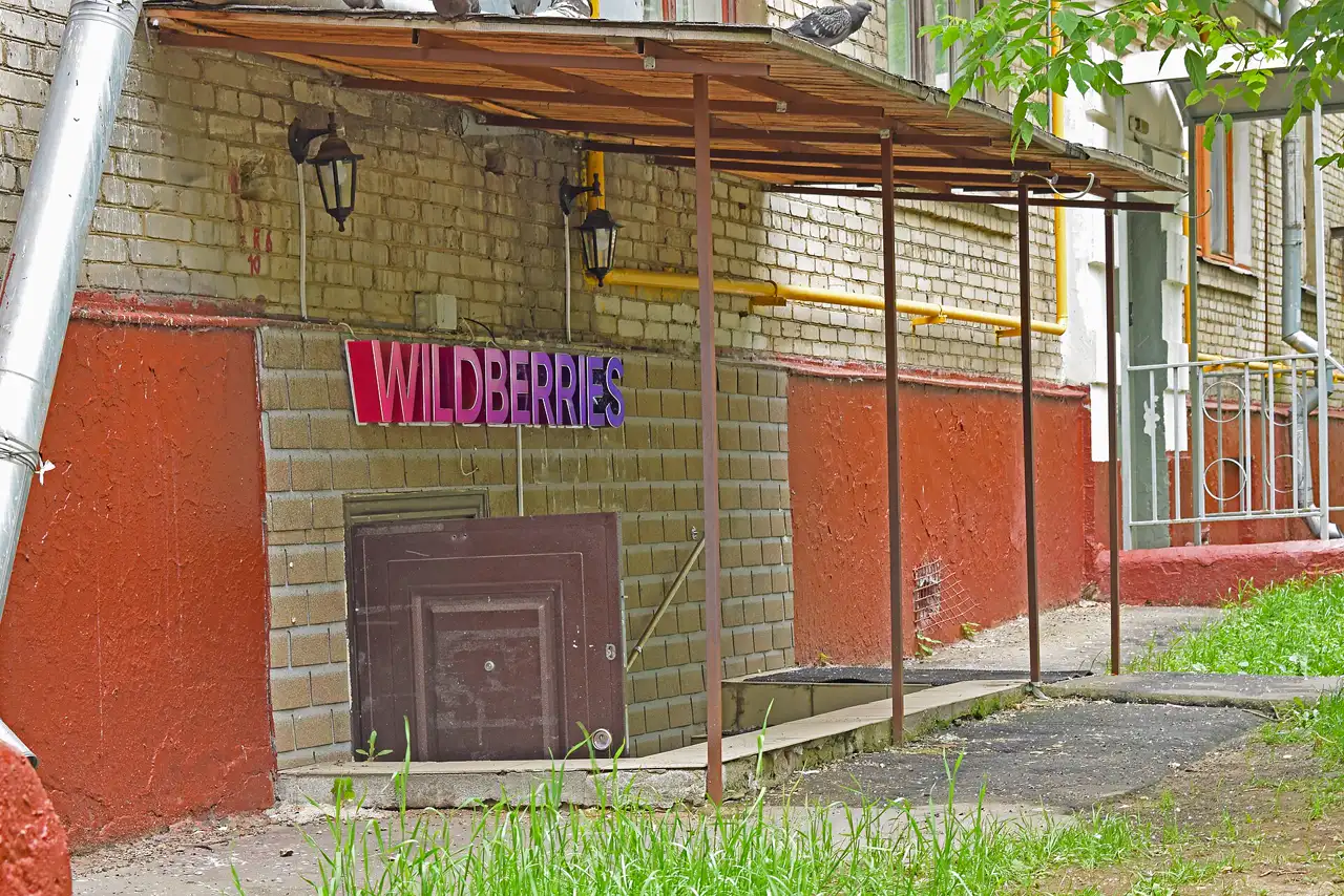 Rótulo de plástico con la palabra WILDBERRIES en degradado de rosa a morado, atornillado a la pared de ladrillo de un edificio; debajo, la puerta metálica de un semisótano y una marquesina de chapa sostenida por postes oxidados.