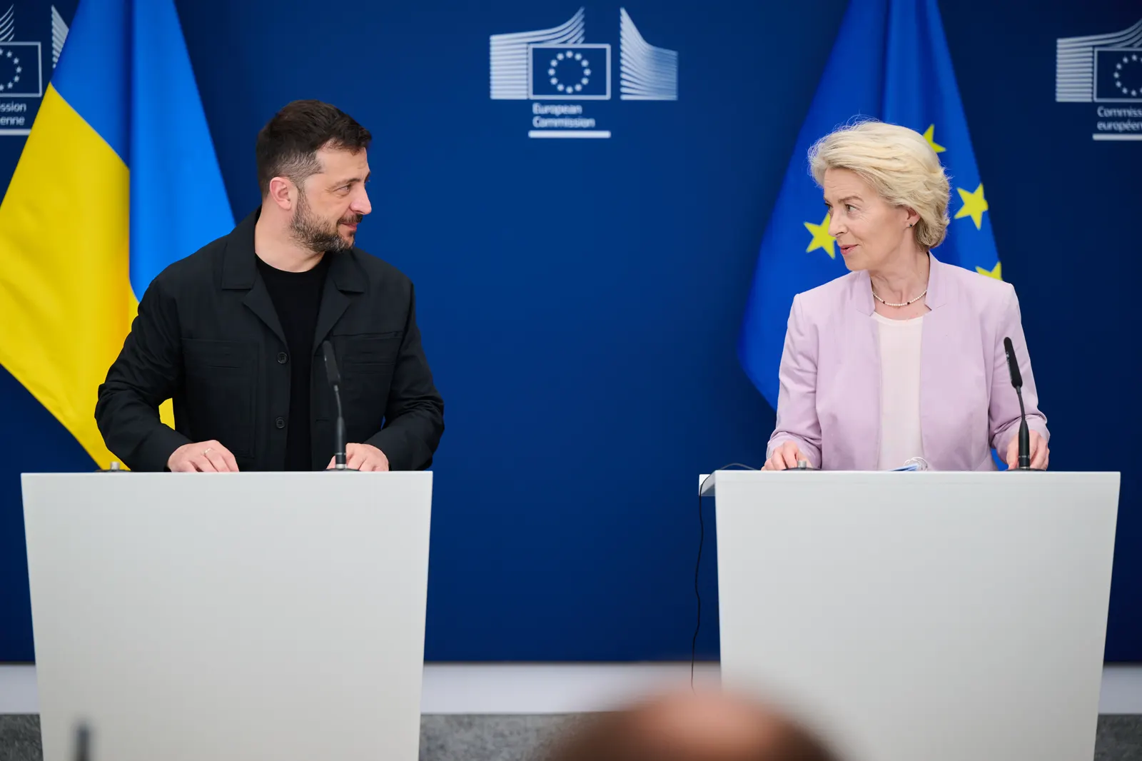 Volodímir Zelenski y Ursula von der Leyen, cada uno tras un atril blanco, se miran durante una comparecencia ante la prensa en la sede de la Comisión Europea; al fondo, un panel azul con el logotipo de la Comisión y banderas de Ucrania y de la Unión Europea.