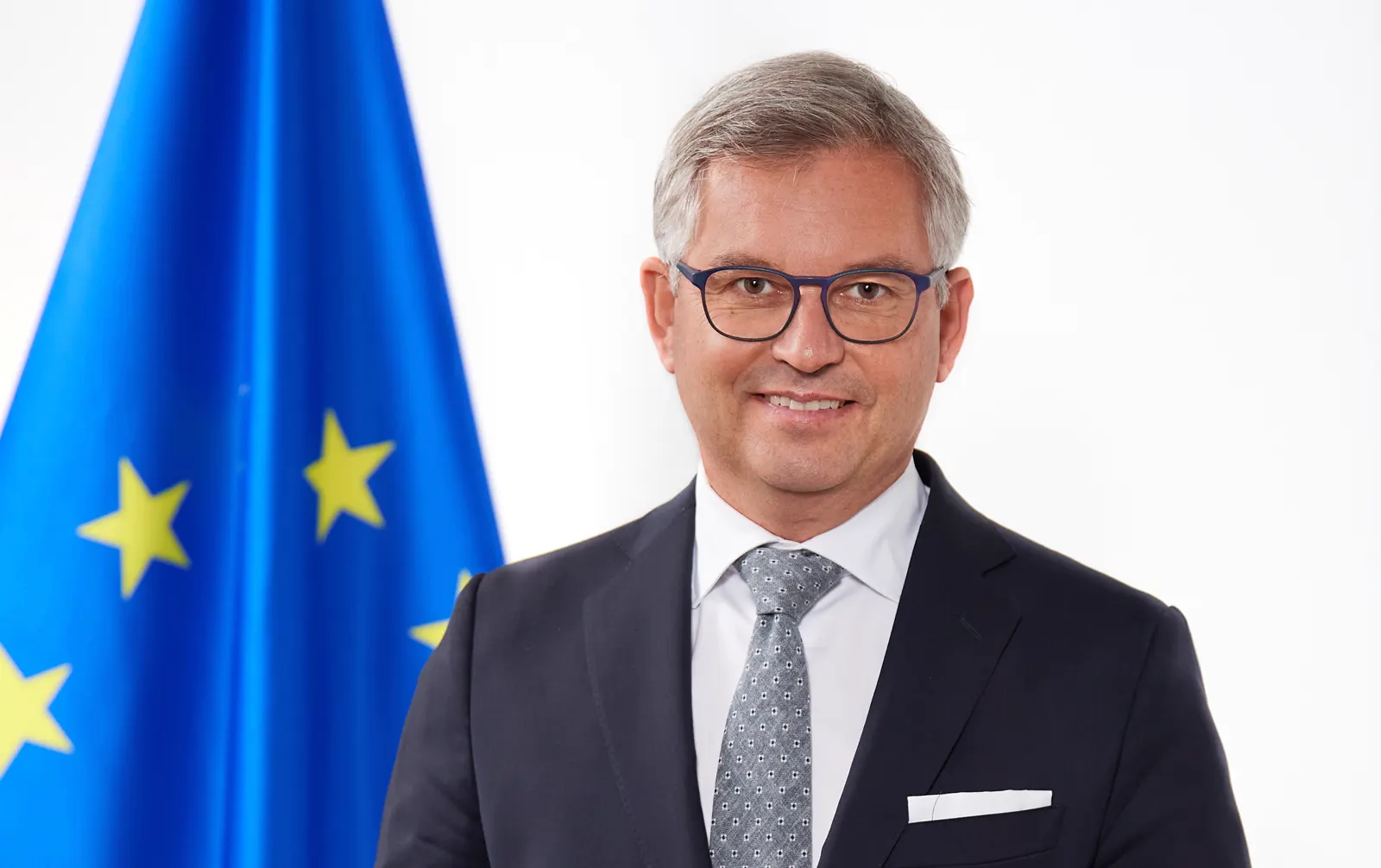 Retrato oficial de Magnus Brunner, comisario europeo de Interior y Migración: hombre de pelo canoso con gafas y traje azul oscuro, sonriente, ante una bandera de la Unión Europea y un fondo blanco.