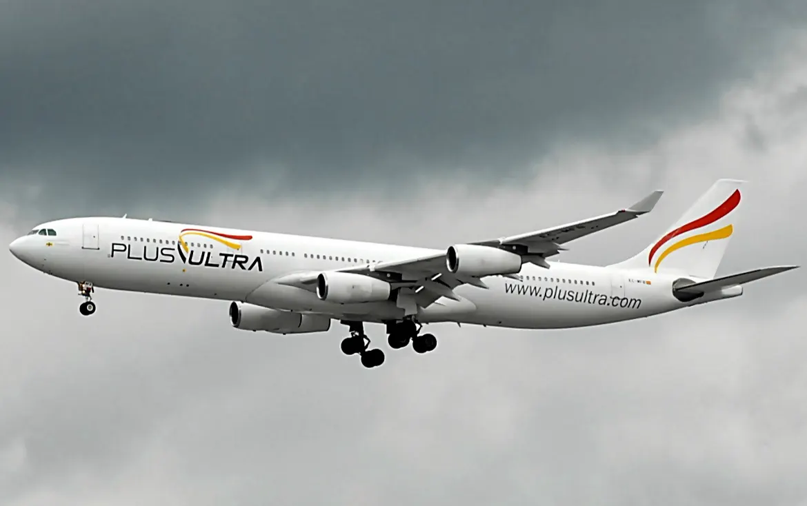 Airbus A340 blanco de Plus Ultra Líneas Aéreas en vuelo, con el tren de aterrizaje desplegado y la dirección plusultra.com rotulada en el fuselaje, bajo un cielo encapotado.