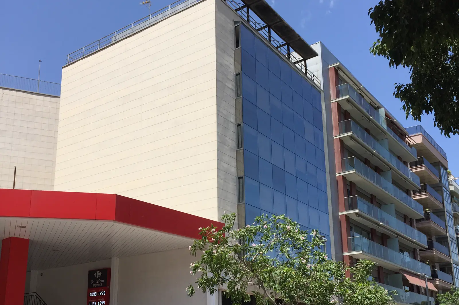 Edificio de oficinas con fachada acristalada azul y lateral de piedra clara, entre una gasolinera y bloques de viviendas, bajo un cielo despejado