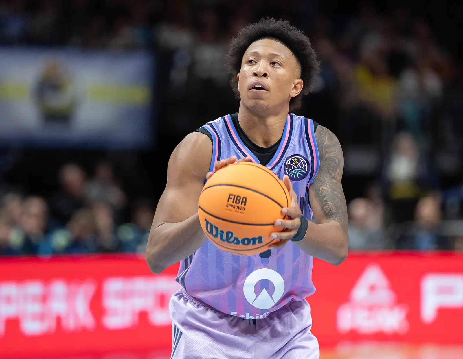 Boogie Ellis, con la camiseta lila del ALBA Berlín, sujeta el balón con las dos manos a la altura del pecho y mira hacia el aro preparado para lanzar, con el público desenfocado al fondo.