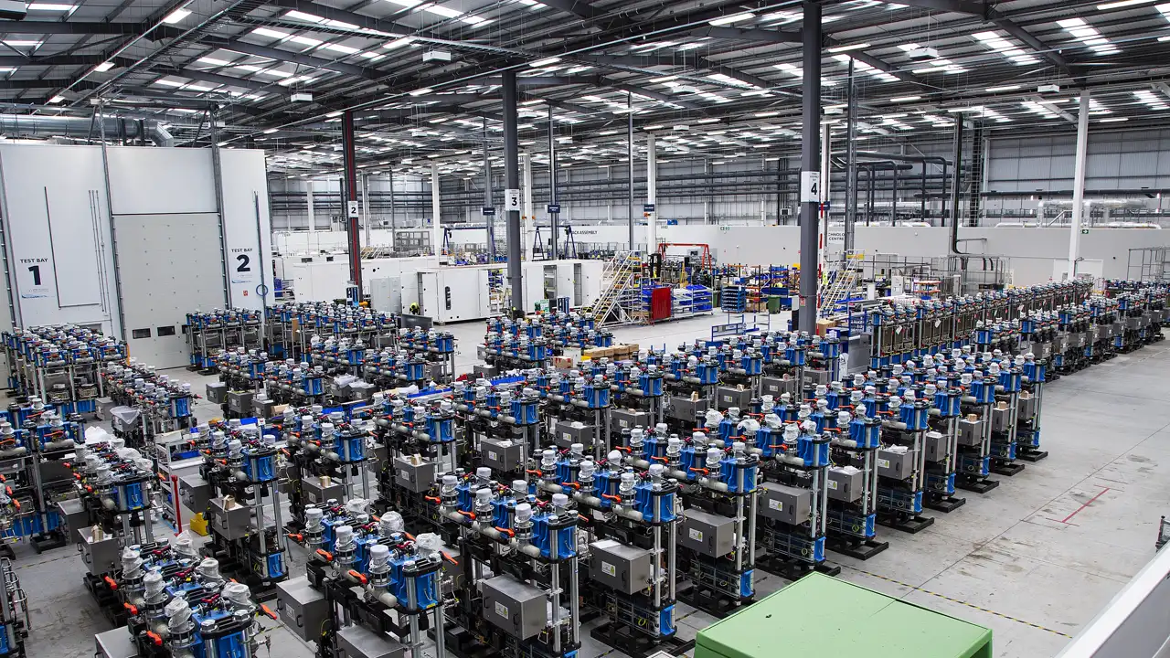 Nave industrial vista desde alto con centenares de módulos de electrolizador alineados en filas, cada uno con una columna azul y depósitos metálicos, junto a dos cabinas blancas de ensayo numeradas.