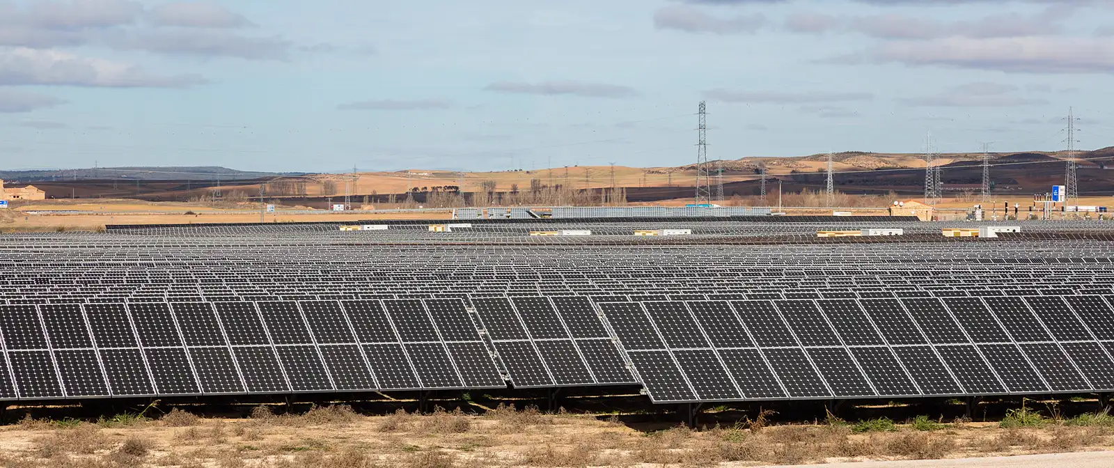 Extensión de paneles solares fotovoltaicos alineados en filas sobre terreno llano y seco; al fondo, varias torres de alta tensión y campos de cereal bajo un cielo con nubes