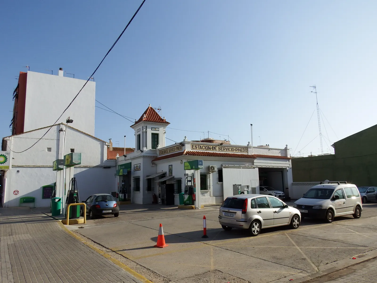 Explanada de una estación de servicio con dos surtidores bajo una marquesina, un rótulo que dice «Estación de Servicio Pinedo» sobre un edificio blanco con tejado de teja, coches aparcados y un cono de tráfico en primer término.