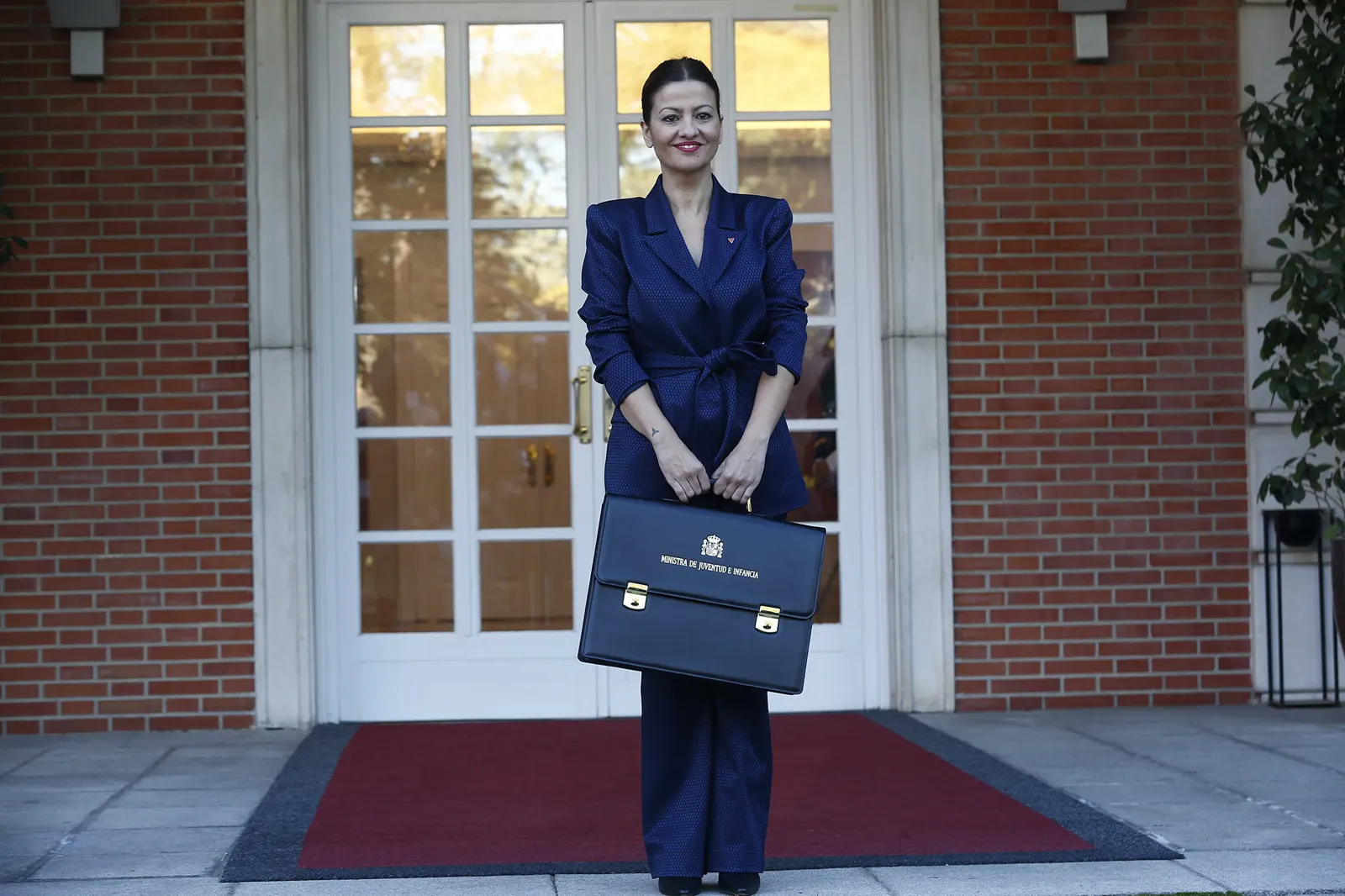 La ministra Sira Rego, de traje azul oscuro, posa de pie ante una puerta acristalada de madera blanca en una fachada de ladrillo rojo, sosteniendo con las dos manos una cartera ministerial negra en la que se lee «Ministra de Juventud e Infancia» bajo el escudo de España.