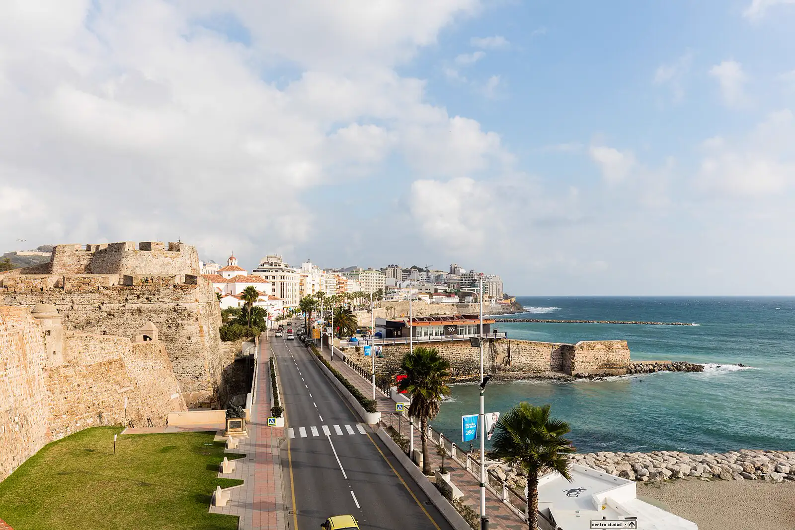 Vista elevada de la costa de Ceuta: a la izquierda, las murallas reales de piedra y una avenida junto al mar; al fondo, los edificios blancos de la ciudad; a la derecha, el Mediterráneo con un espigón de rocas y una playa.