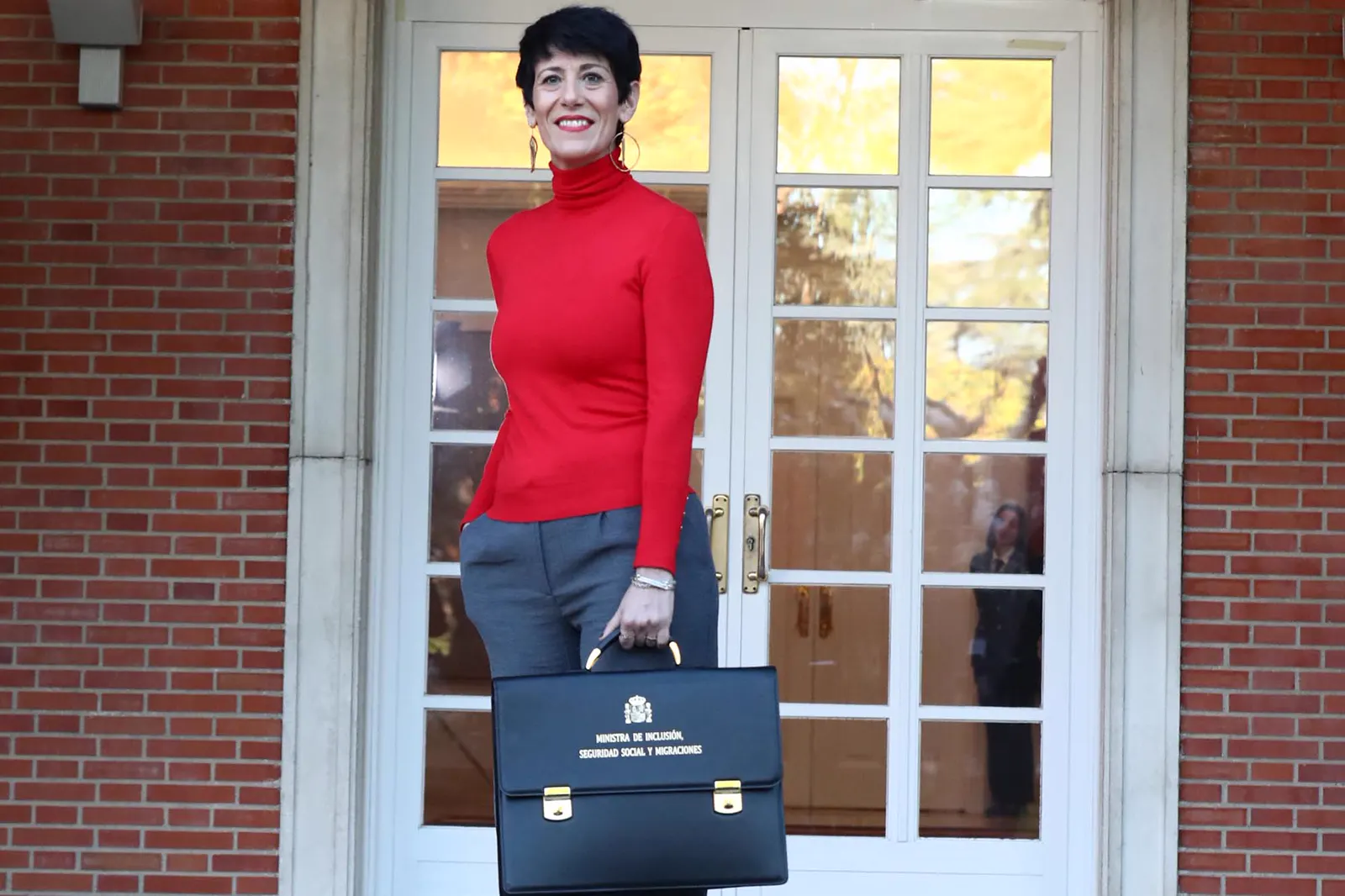 Elma Saiz, con jersey rojo de cuello alto y pantalón gris, posa de pie ante una puerta acristalada blanca en una fachada de ladrillo rojo, sosteniendo con una mano una cartera ministerial negra en la que se lee «Ministra de Inclusión, Seguridad Social y Migraciones» bajo el escudo de España.