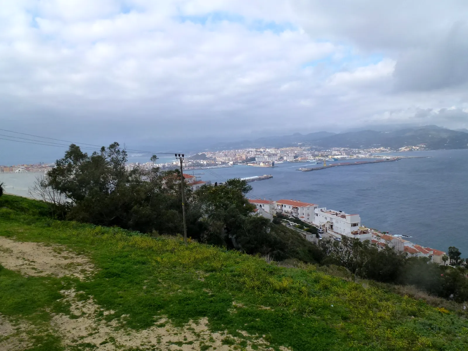 Vista de Ceuta desde una ladera del Monte Hacho: en primer término, una loma de hierba con un poste eléctrico; a la derecha, casas blancas de tejado rojo escalonadas sobre el mar; al fondo, el caserío de la ciudad, los diques del puerto y las montañas bajo un cielo nublado.