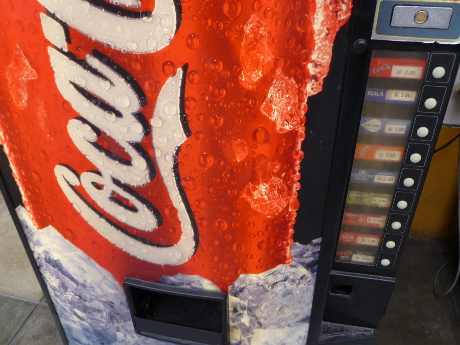 Máquina expendedora roja de Coca-Cola con botellas y latas visibles tras el cristal, en una calle de Lima.