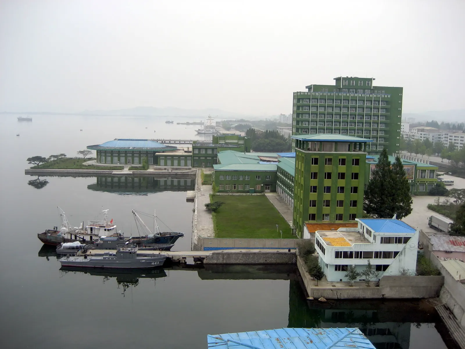 Vista elevada de la bahía de Wonsan en un día brumoso: edificios verdes de tejado azul al borde del agua, varios pesqueros y una lancha amarrados al muelle y un carguero difuminado en el horizonte.