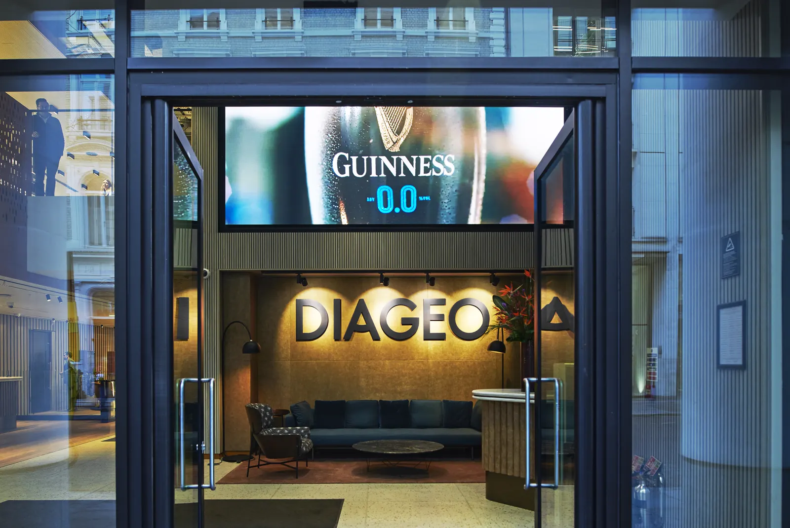 Entrada acristalada de la sede mundial de Diageo: al fondo, el rótulo DIAGEO iluminado sobre la recepción y una pantalla con un anuncio de Guinness 0.0