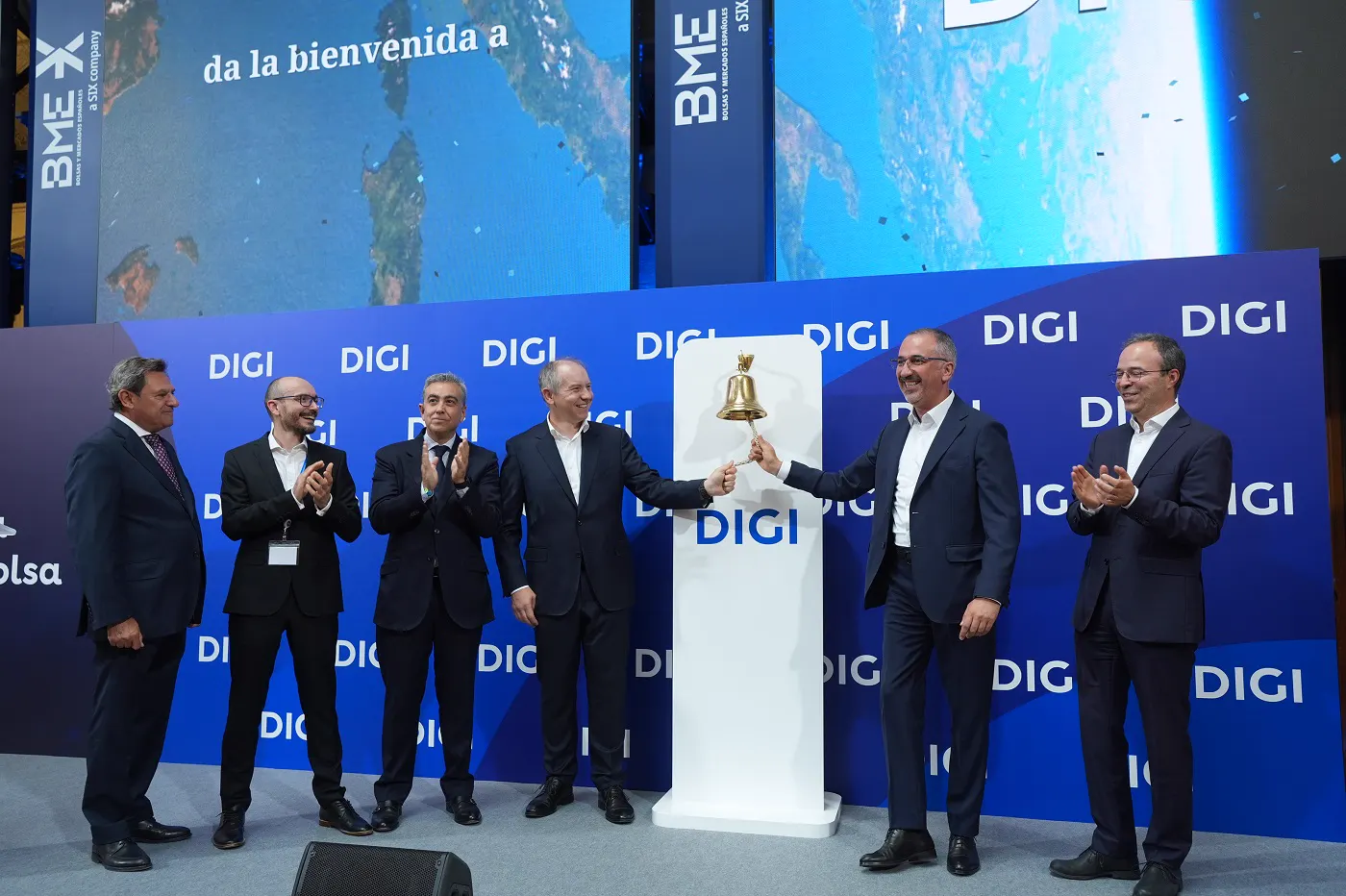 Seis directivos trajeados, dos de ellos tocando una campana dorada sobre un pedestal con el logotipo de DIGI, en un escenario con paneles de DIGI y de BME durante el estreno bursátil de la compañía.