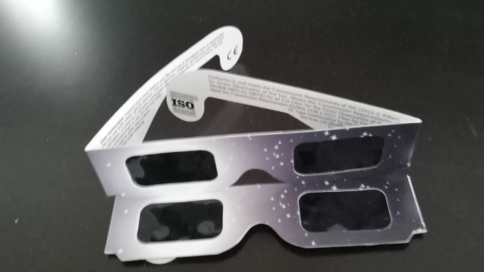 Gafas de eclipse de cartón plateado con motivos de estrellas, con los cristales negros opacos y el sello ISO y el marcado CE impresos en la varilla, sobre una superficie oscura