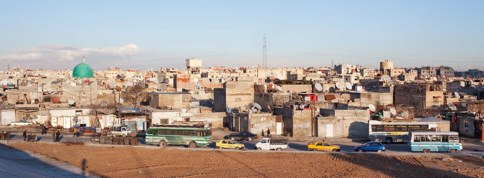 Vista general de los tejados y las casas bajas de la localidad de Yaramana, a las afueras de Damasco, con varios autobuses, minibuses y taxis circulando por una avenida en primer plano