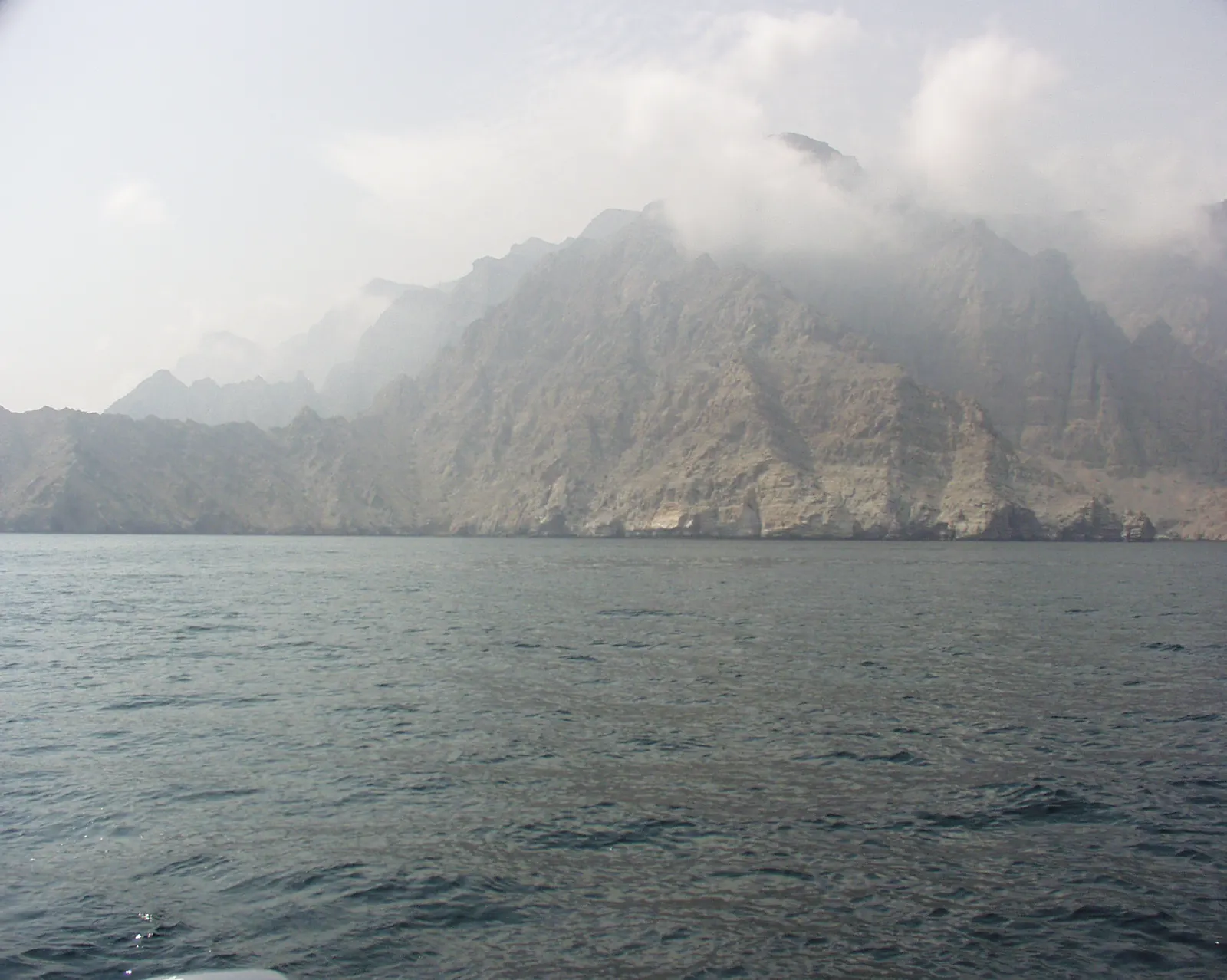 Acantilados rocosos y montañas áridas envueltas en bruma se levantan sobre un mar gris azulado, vistos desde una embarcación.