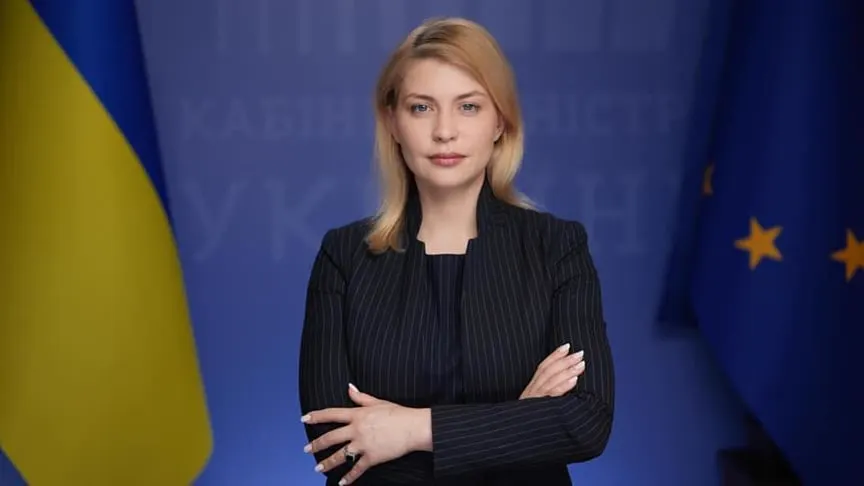 Mujer rubia con traje oscuro de raya diplomática, de pie y con los brazos cruzados, ante un panel azul con la bandera de Ucrania a un lado y la de la Unión Europea al otro.