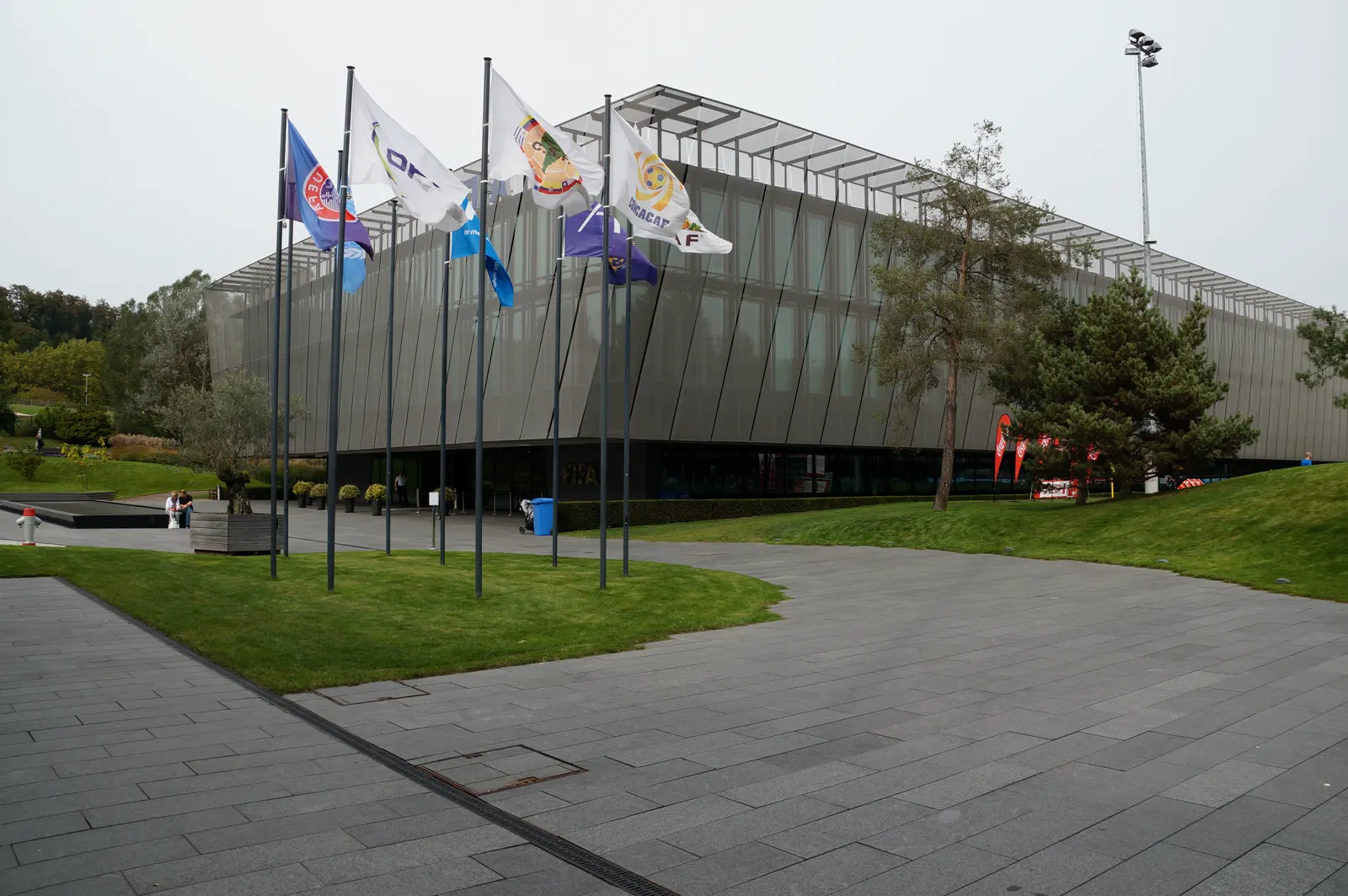 Edificio acristalado y de perfil quebrado de la sede de la FIFA en Zúrich, con una hilera de banderas de confederaciones y federaciones en mástiles sobre una explanada empedrada