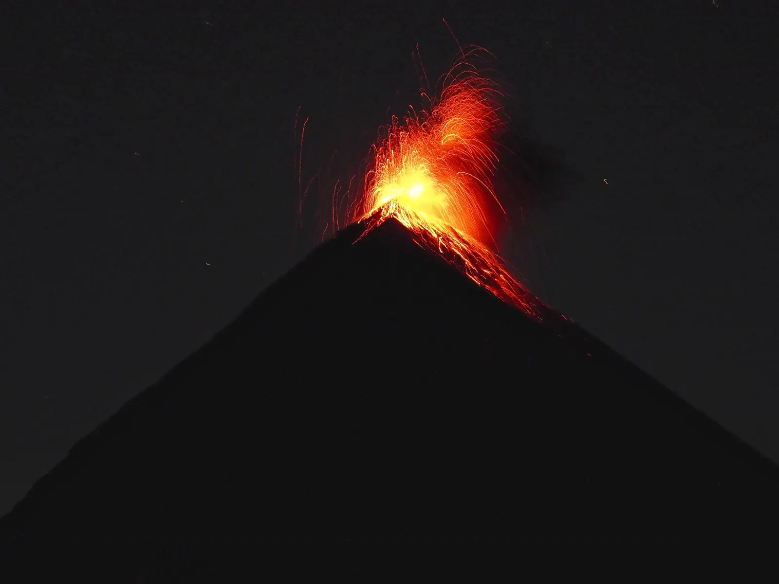 Silueta del cono del volcán de Fuego de noche, con una fuente de lava incandescente saliendo del cráter y material ardiente rodando por la ladera derecha