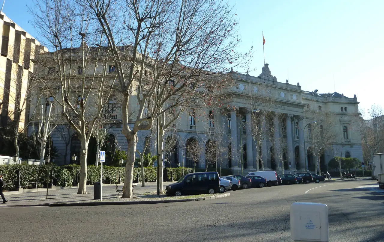 Fachada de piedra del Palacio de la Bolsa de Madrid, con columnas y reloj en el frontón, tras una hilera de plátanos sin hojas y coches aparcados en la acera.