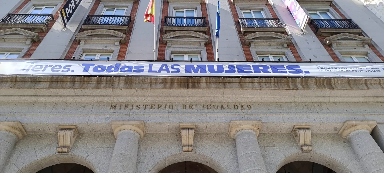Fachada de un edificio de ladrillo y piedra con la inscripción «Ministerio de Igualdad» tallada sobre un soportal de columnas, con las banderas de España y de Europa y una pancarta institucional colgada de los balcones.