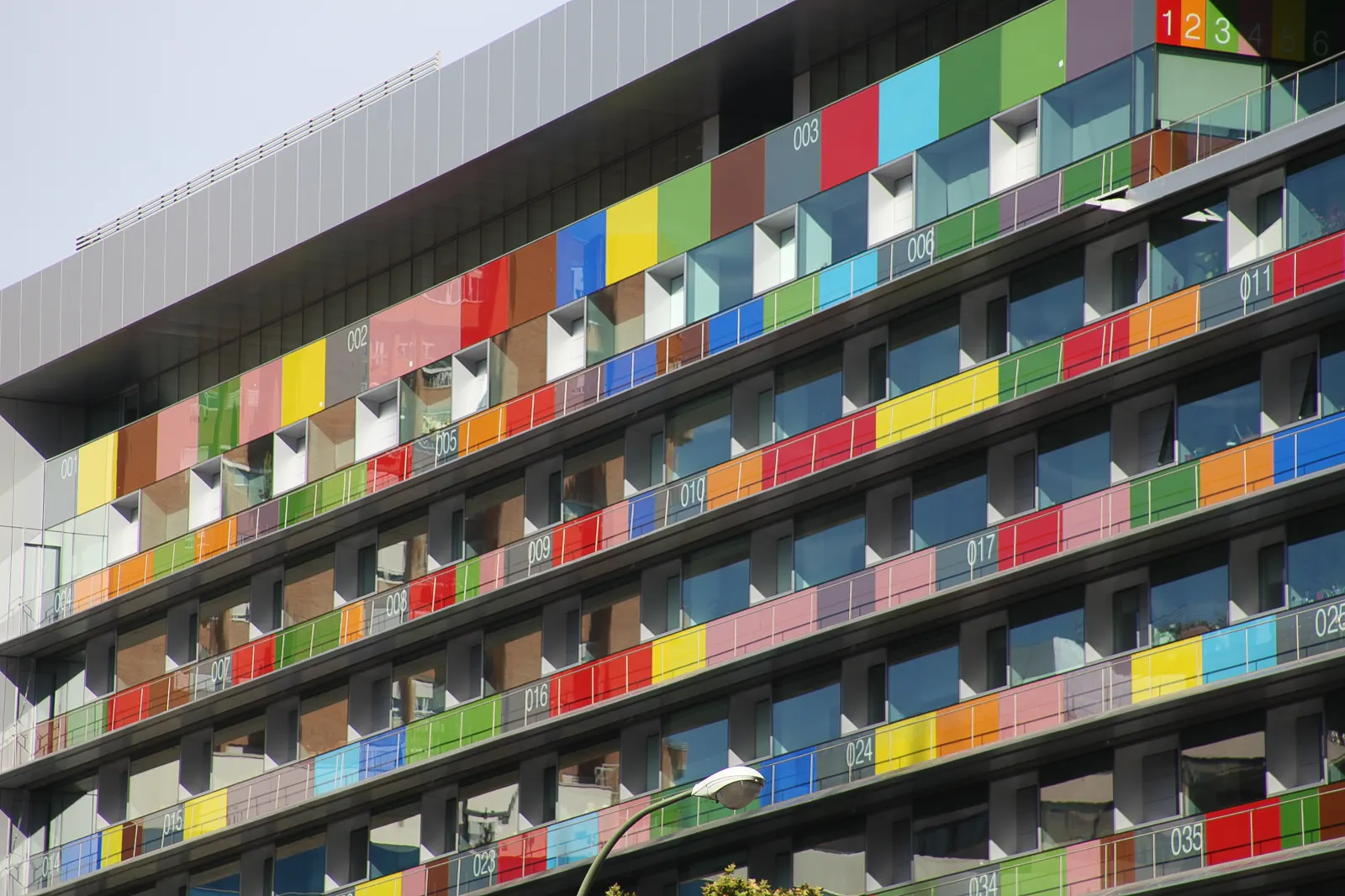 Detalle de la fachada del edificio del Instituto Nacional de Estadística en Madrid: hileras de paneles de vidrio de colores —rojos, azules, verdes, amarillos— con números impresos en blanco sobre cada balcón.