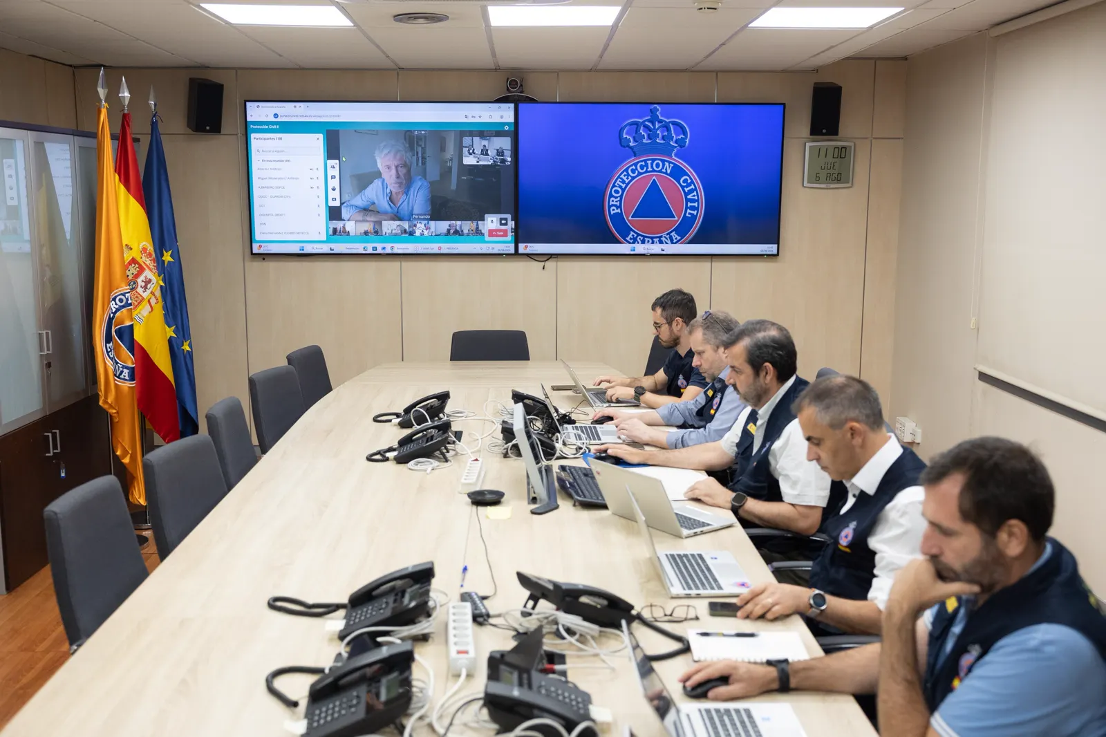 Sala de reuniones con banderas de España y de la Unión Europea y una bandera con el emblema de Protección Civil; en dos pantallas de pared, una videoconferencia con un participante rotulado «Fernando» y el escudo de Protección Civil de España. Cinco hombres con chalecos y polos de Protección Civil trabajan con ordenadores portátiles a lo largo de una mesa alargada llena de teléfonos. Un reloj de pared marca las 11.00 del jueves 6 de agosto.