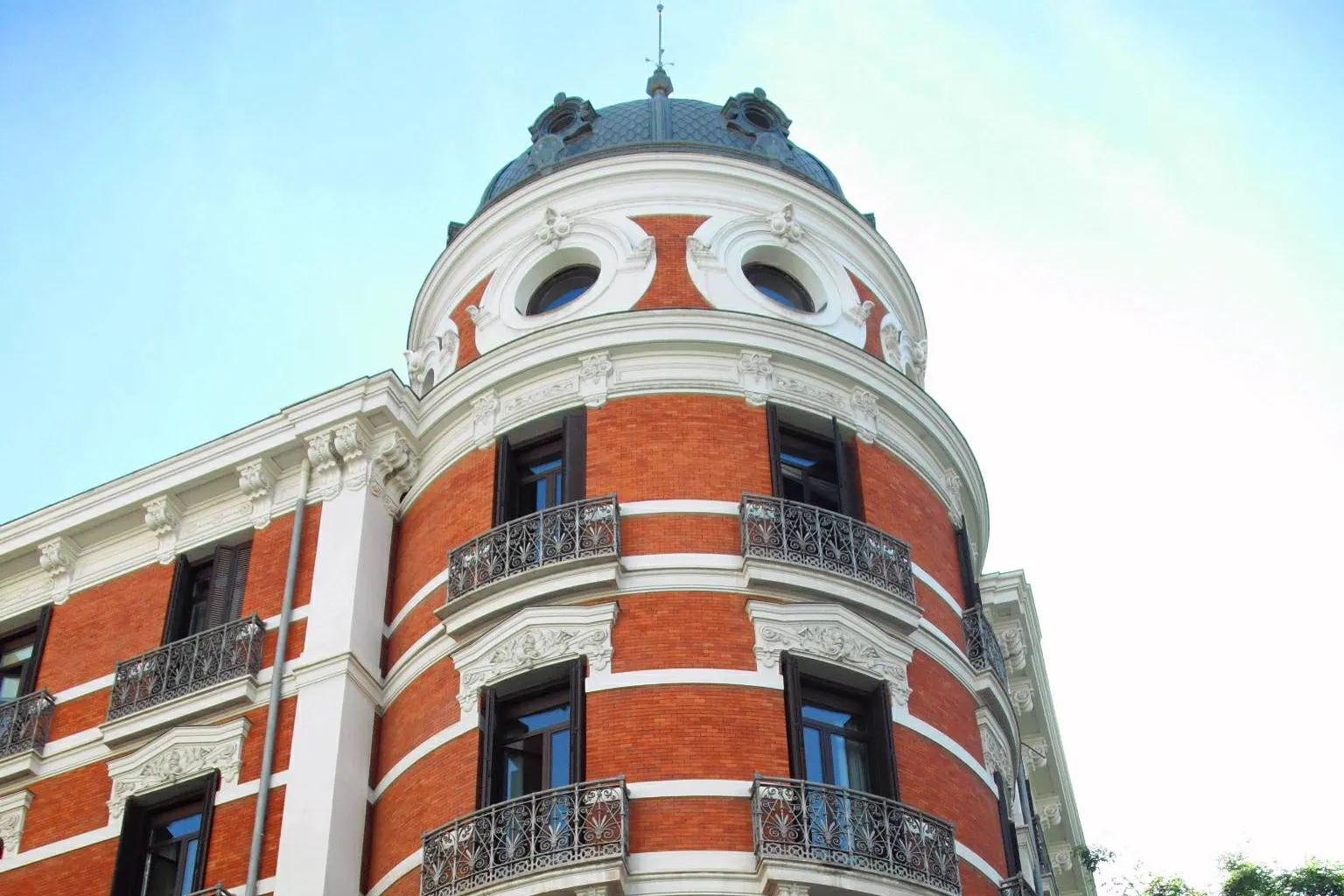 Esquina curva de un edificio de ladrillo rojo y molduras blancas rematada por una cúpula de pizarra azulada con dos óculos, con balcones de hierro forjado y el cielo despejado al fondo.