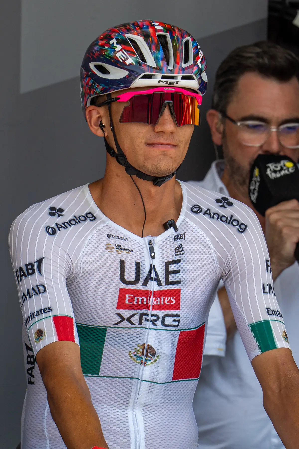Isaac del Toro, con casco y gafas de sol, vestido con un maillot blanco del UAE Team Emirates-XRG cruzado por franjas verde y roja y el escudo de la bandera de México, mira al frente antes de la salida de una etapa.